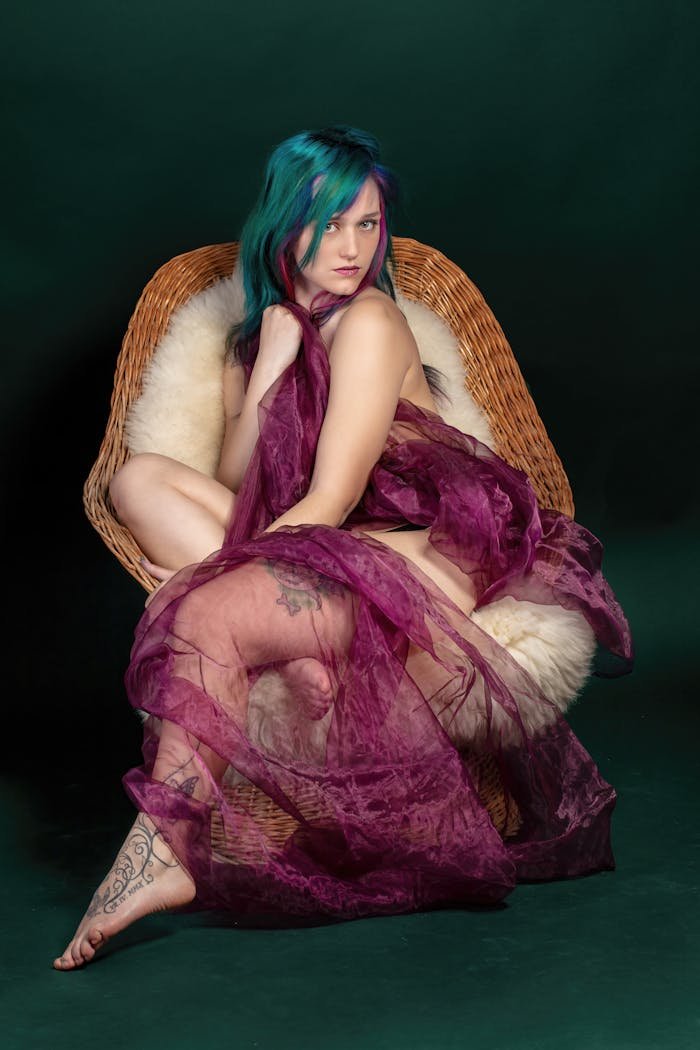 who-we-are Portrait of a young woman with vibrant hair posing elegantly with translucent fabric in a wicker chair.