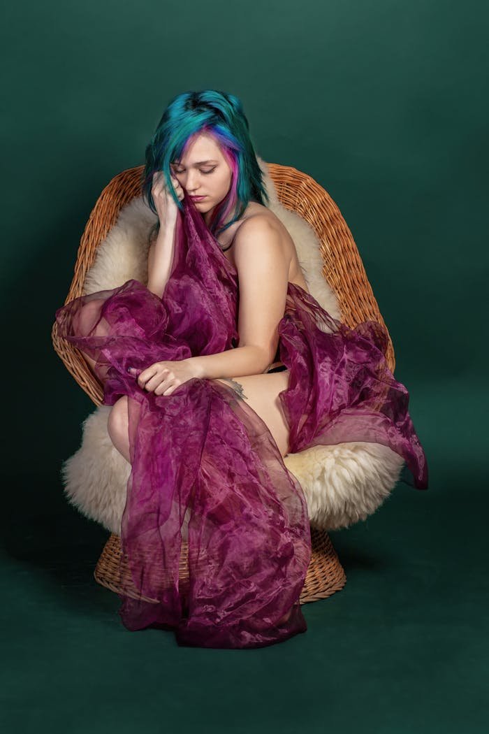 our-story Artistic portrait of a woman with colorful hair in a fashion-forward pose, draped in purple fabric.